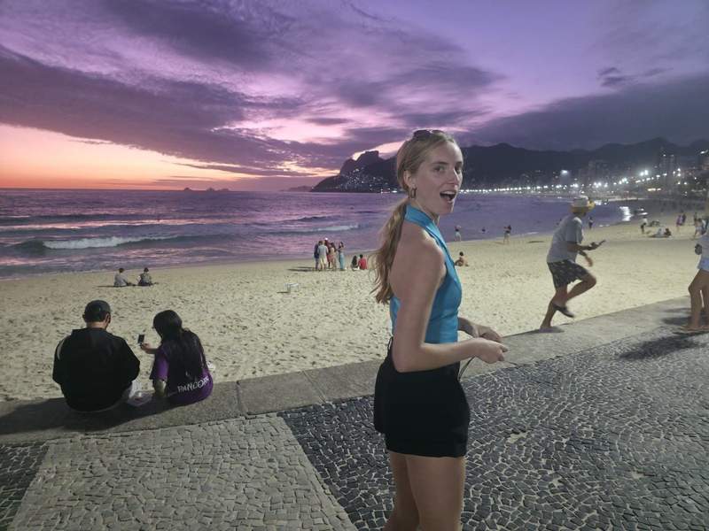 Author standing along the Copacabana promenade at dusk with the ocean beachgoers and Rios illuminated coastline stretching behind her under a purple sunset sky.