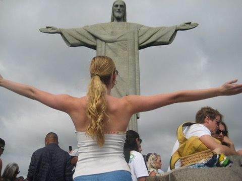Author standing with arms outstretched facing the Christ the Redeemer statue in Rio de Janeiro with other visitors nearby on a cloudy day.
