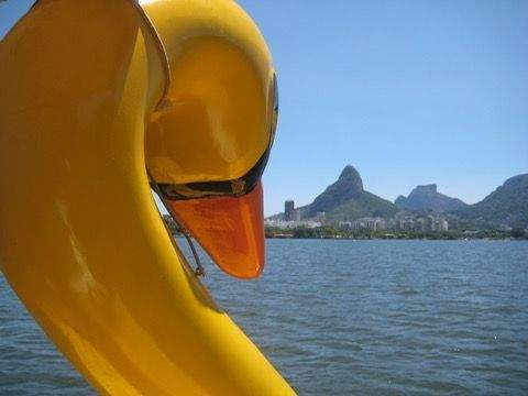 Yellow duck shaped pedal boat on Lagoa Rodrigo de Freitas in Rio de Janeiro with calm water and mountain silhouettes in the background under a clear blue sky.