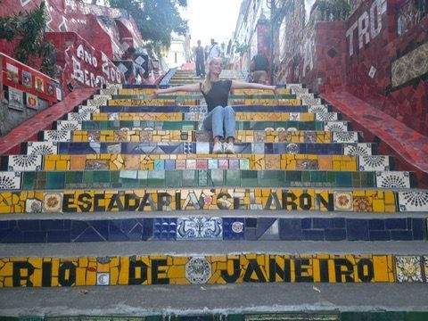 Authir seated on the brightly tiled Escadaria Selaron staircase in Rio de Janeiro surrounded by colorful mosaic tiles and street art with people walking above.