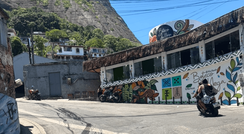 Colorful favela in Rio de Janeiro with a vibrant hand painted mural motorcycles passing by and lush green mountains in the background.
