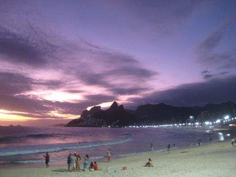 Beach at sunset in Rio de Janeiro with warm orange and pink skies calm waves and people gathered along the shoreline.