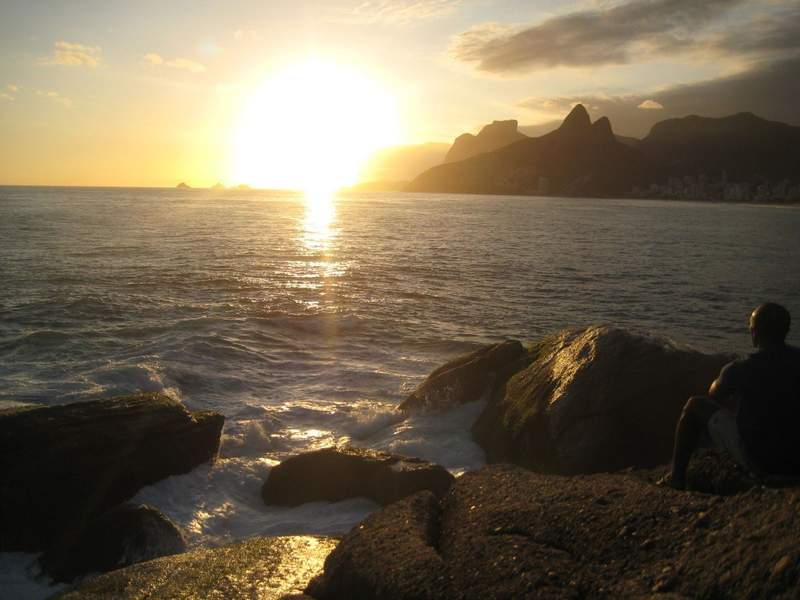 Golden sunset over the ocean in Rio de Janeiro with waves breaking against dark rocks in the foreground and mountain silhouettes lining the coastline in the background.