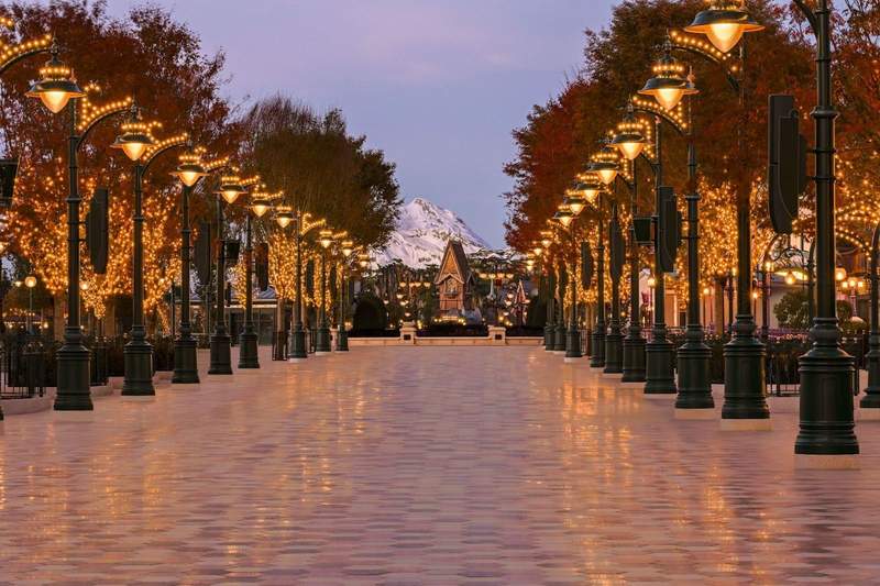 An inviting tree lined promenade is lit by decorative streetlamps at Disneyland Paris Disney Adventure World. The Kingdom of Arendelles village clocktower can be seen in the distance. 
