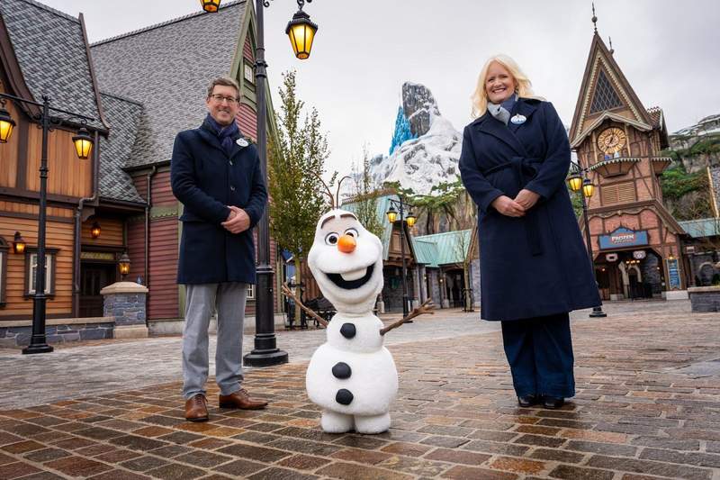 Disneyland Paris executives pose with new free roaming Audio Animatronic figure of Olaf surrounded by brightly painted Norwegian building with the village clocktower behind them.