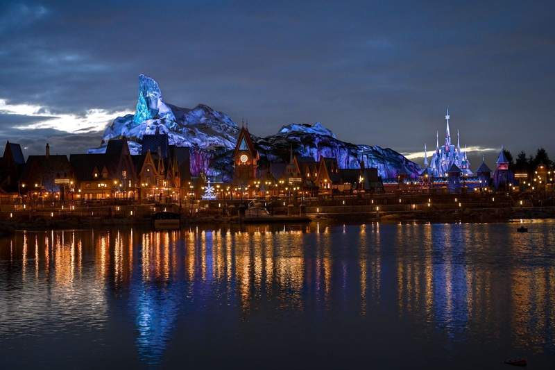 The glittering skyline of Disneyland Paris World of Frozen at night as seen from across the Bay of Arendelle. The village lights reflect across the water making it appear to glow. 