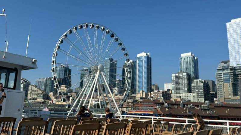 wooden chairs on a boat with a large ferris wheel in the distance against skyscrapers of varying sizes in Seattle.