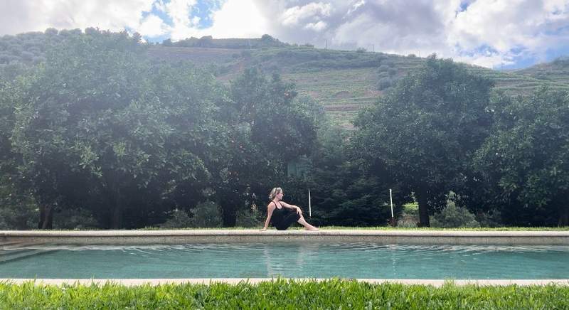 A woman in black sits on the side of a pool surrounded by green foliage.