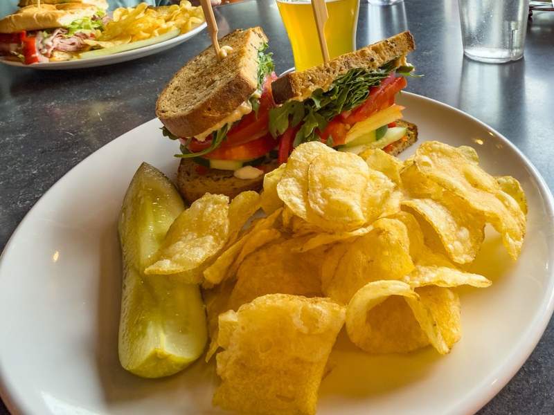 White plate piled high with a sandwich potatoe chips and dill pickle with a beer in the background.