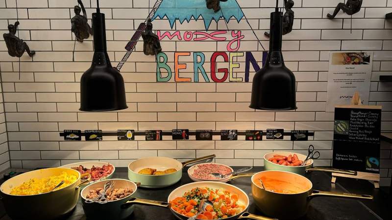 Sausage eggs and vegetables sitting inside of green pots on a stove with a Moxy Bergen illustration on the tile wall. 