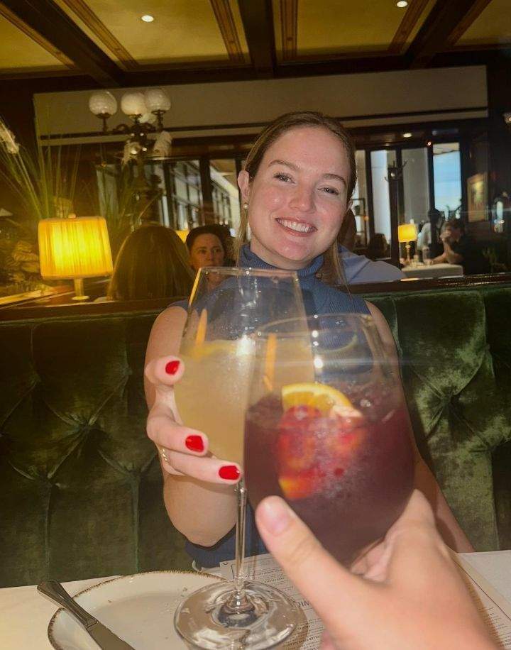 A woman joyfully toasts with a cocktail in a bustling restaurant enjoying the timeless vibe of Old Ebbitt Grill with her friend.