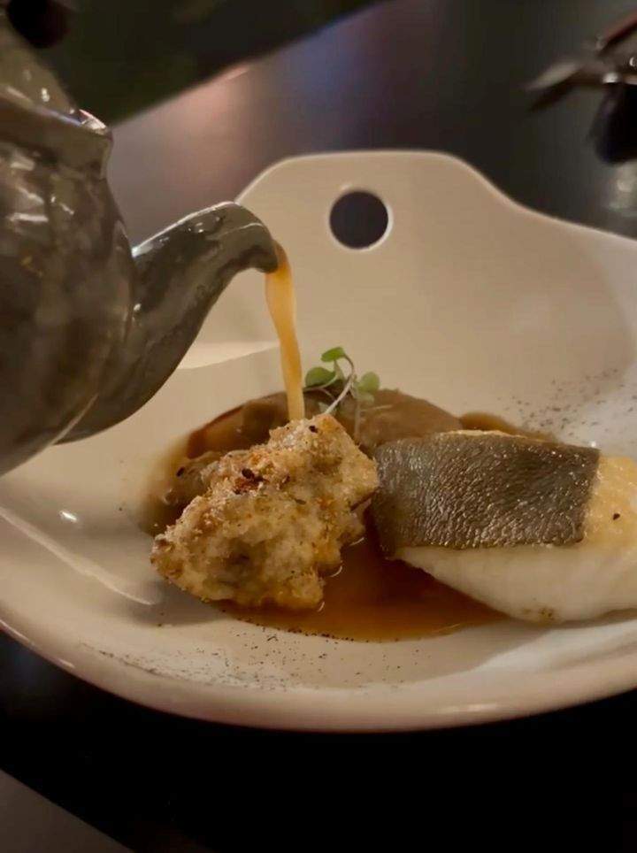  A white plate with an artistic dish of a fish dish from Cranes 7 Course Omakase with a teapot pouring broth.