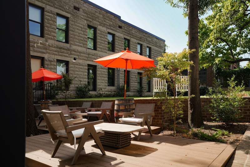 An outdoor patio with orange umbrellas table and chairs sits in a garden next to the hotel. 