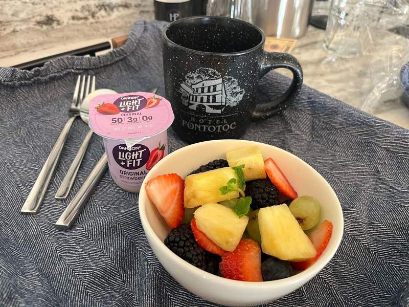 A tray holds silverware a coffee mug yogurt and fresh bowl of fruit.