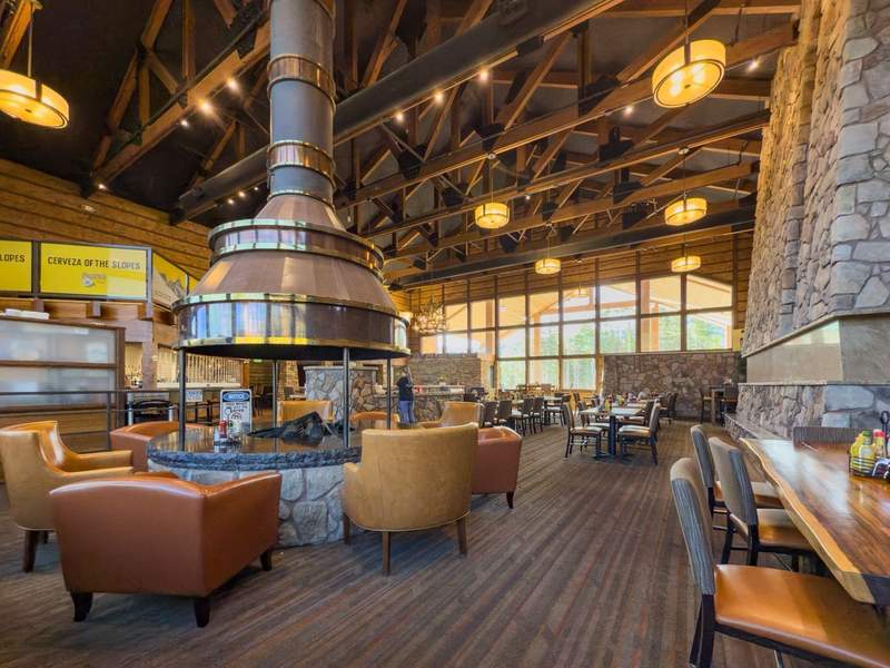 Mountain lodge dining room with open beamed ceiling a round fireplace in the center surrounded by easy chairs.