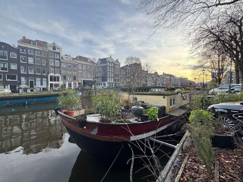 A fully lived in houseboat parked in a canal in Amsterdam silhouetted by a yellow sunset.