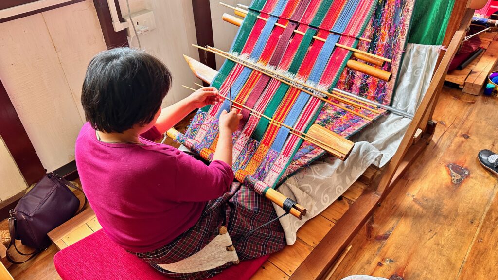 A woman with short, dark hair sits at a floor loom with brightly-colored yarns.