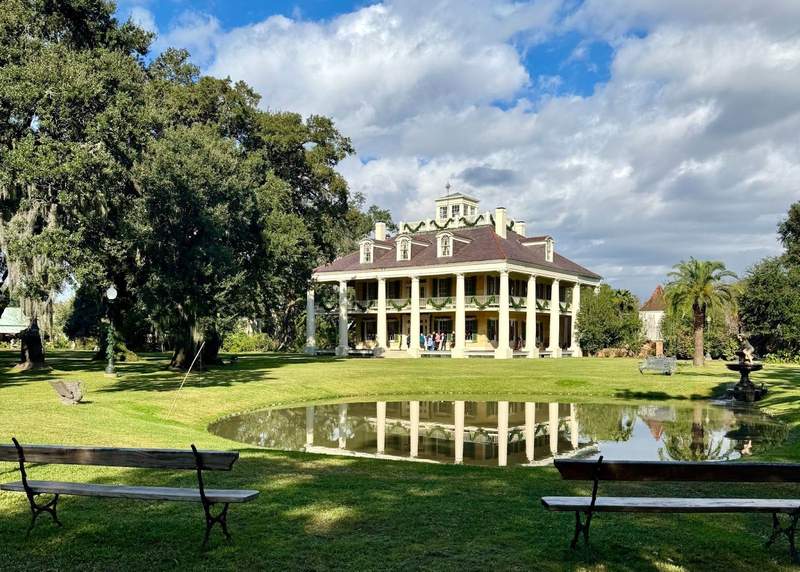 A white house with columns is decorated with garlands for Christmas and reflected in a small lake