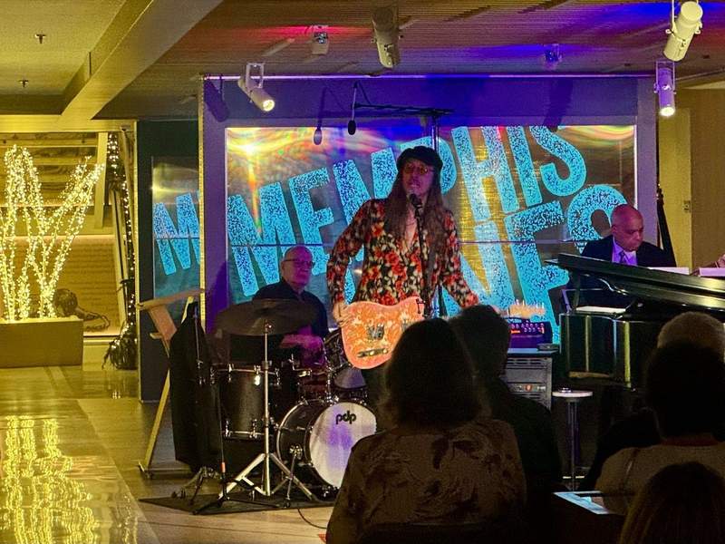 Man in flowery shirt and hat sings in microphone with band behind him
