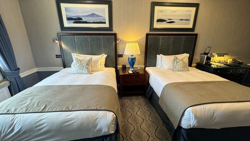 two full beds with grey runners beneath pieces of artwork with a night lamp in between next to a desk at The Shelbourne