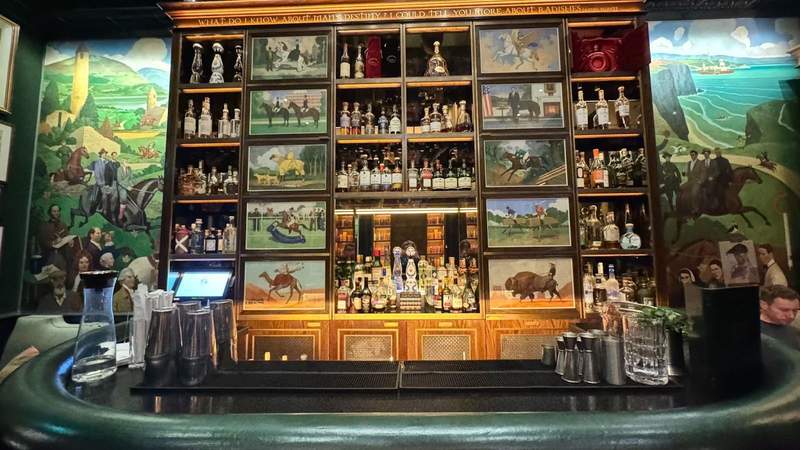 half circle bar in front of small murals of horses and camels next to glass bottles of alcohol from the 1824 bar at The Shelbourne.