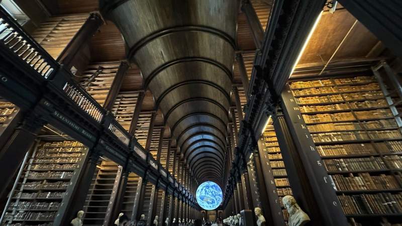 large library with thousands of books on wooden shelves and an arched ceiling with illuminated globe at the end at Trinity College