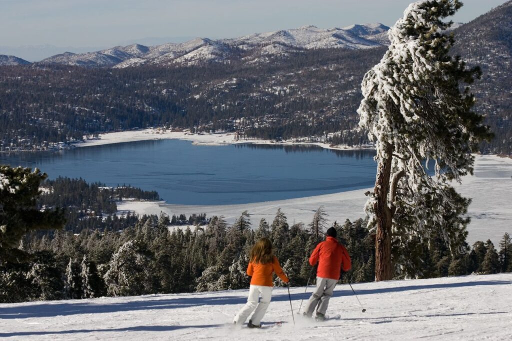 Couple in orange jackets skiing down a slope overlooking Big Bear Lake and snowcapped mountains in the distance.