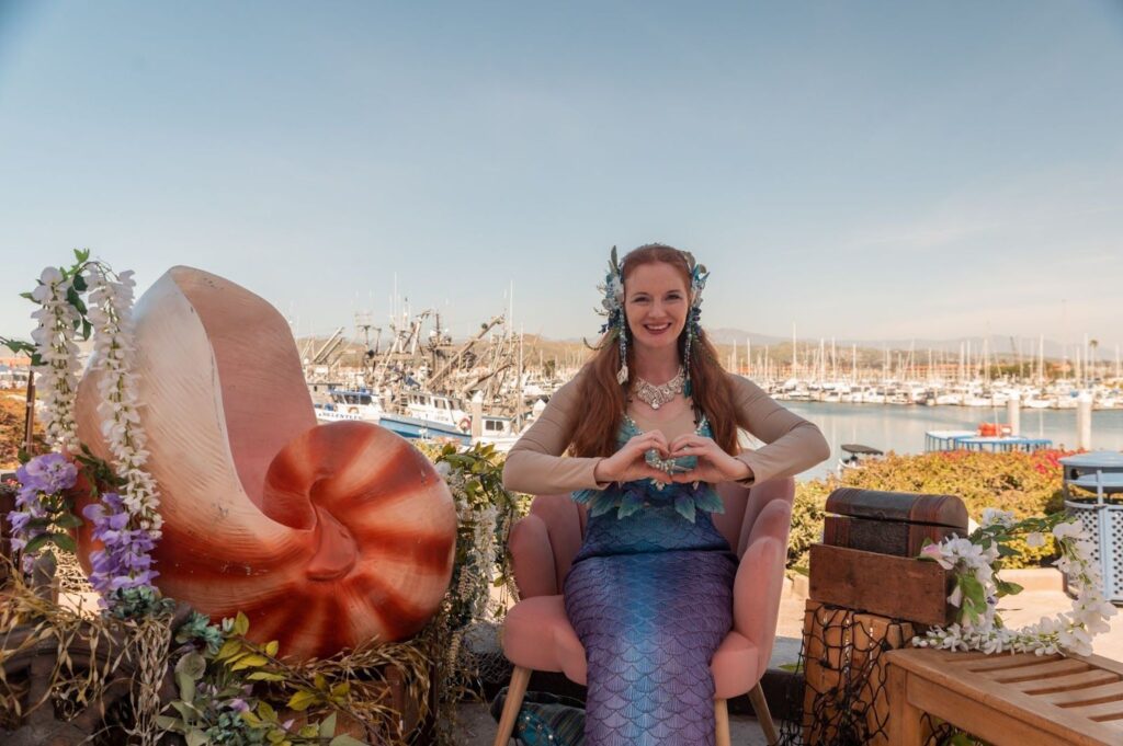 Woman dressed in purple mermaid costume next to a large shell overlooking the marina at Ventura Harbor Village.