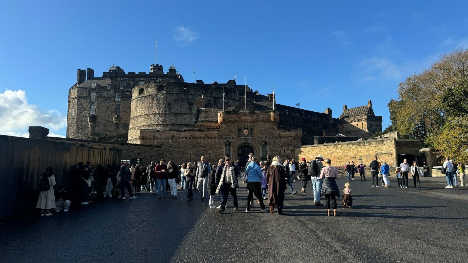 People walking on pavement outside of Edinburgh Castle with flag poles standing upright on top of the building.