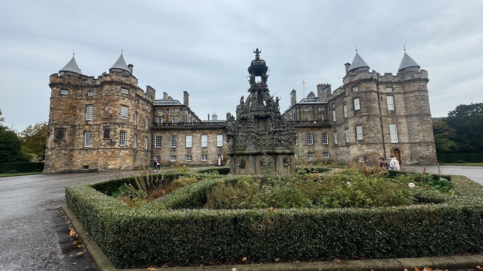 Tall palace building with pointed rooftops and dozens of windows in back of a green shrub around a statue in Edinburgh