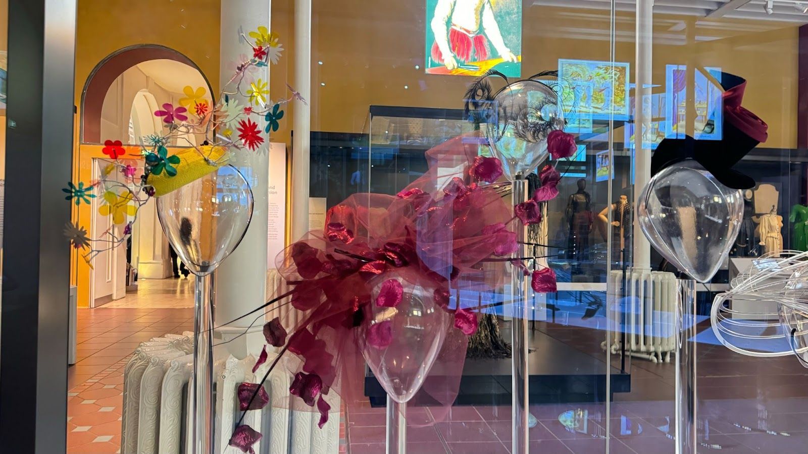 clear mannequin heads in a glass display case with different hats including rainbow floral ones and large pink roses at the National Museum of Scotland