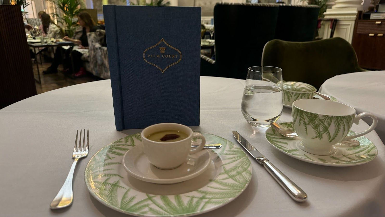 Tea cup on a small plate stacked on a large green plate with palm leaves in front of a blue Palm Court menu at the afternoon tea held at The Balmoral in Edinburgh.
