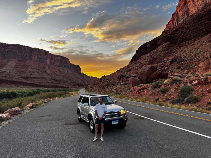 A man stands in front of a silver SUV on a winding desert road at sunset, surrounded by towering red rock canyon walls and glowing clouds overhead.