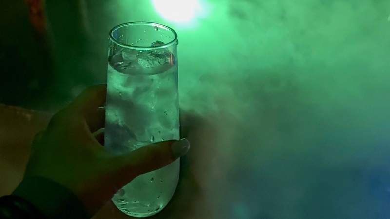 A hand holds a clear drink over ice in a misty, green-lit tasting room during the immersive cocktail experience at Obscure Distillery. Photo credit: Kim Orlando
