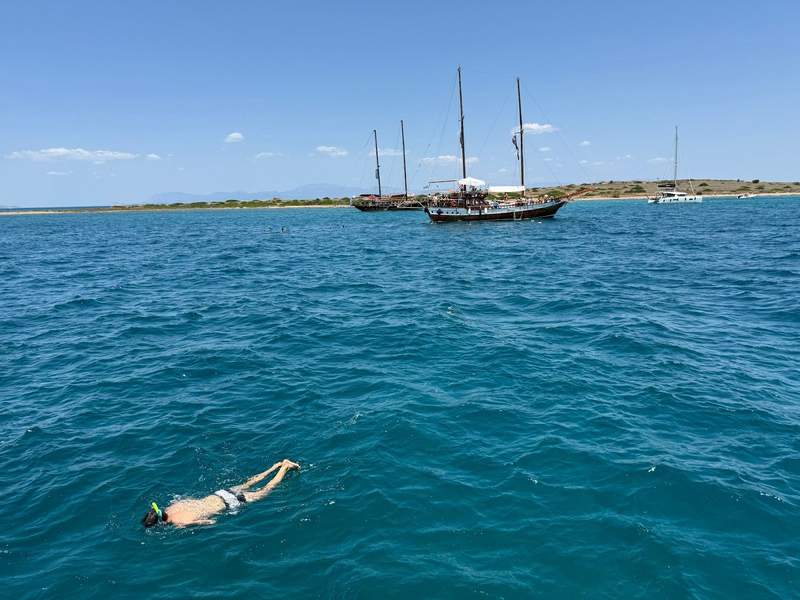 Man snorkeling in blue water with several sailboats and a small Greek island in the background.