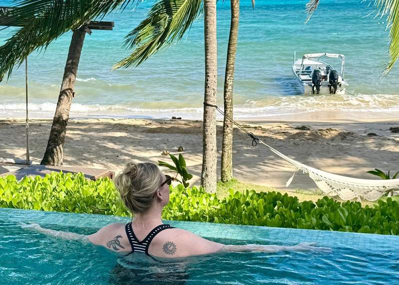 A woman enjoys a dip in the pool while looking out at the ocean and a boat tied up near shore.