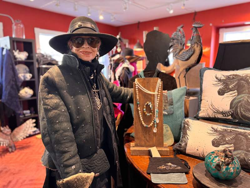 Sallie Bell, fashion designer, wearing a hat, sunglasses and a jacket, standing next to her designs.