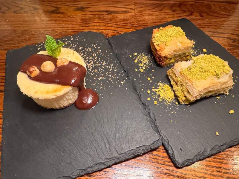 Two black slate tiles with a hazelnut cheesecake with chocolate ganache and pistachio baklava.