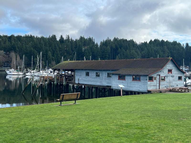 Grassy park overlooking Gig Harbor and a marina filled with boat, historic building known as a netshed.
