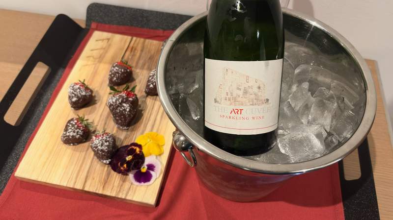 Strawberries covered with chocolate on a wooden plate on the left. A bottle of sparkling wine in an ice bucket on the right.
