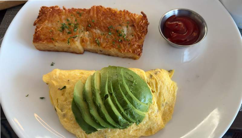 On top, two crispy squares of a potato casserole with a small silver bowl filled with ketchup. In the bottom is a yellow omelet with an avocado sliced and placed on top of it.