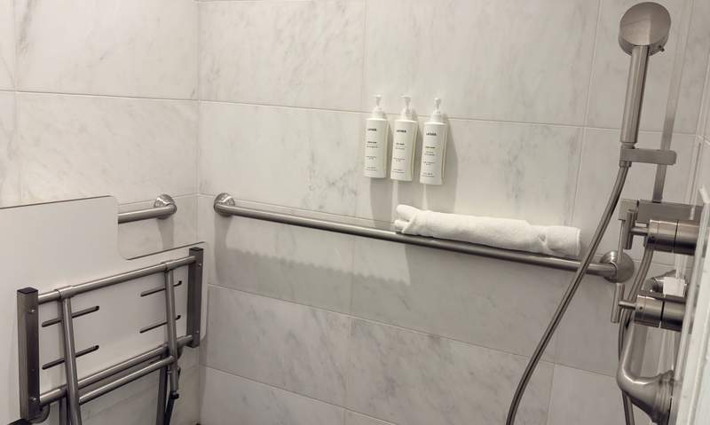 Seat closed against the wall in the shower with rail next to it. The middle has rail plus white plastic bottles of shampoo, conditioner and body wash.