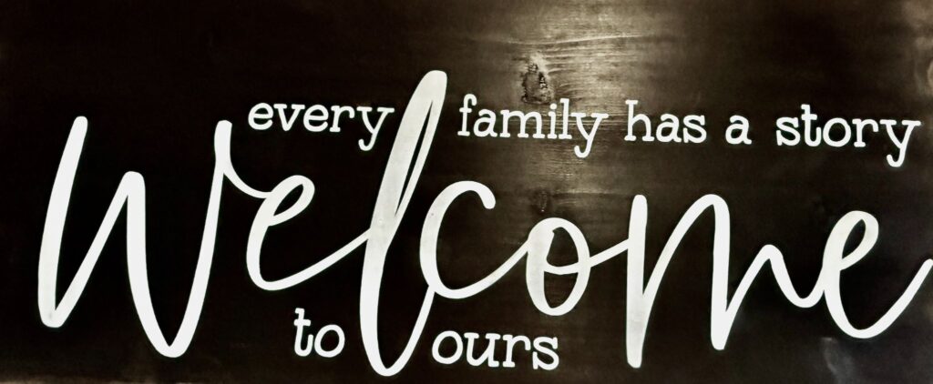 Welcome is the big word on this black and white horizontal sign, written in script. Lover case printed letters declare every family has a story and welcome to ours. The sign hangs in the entrance of a restaurant