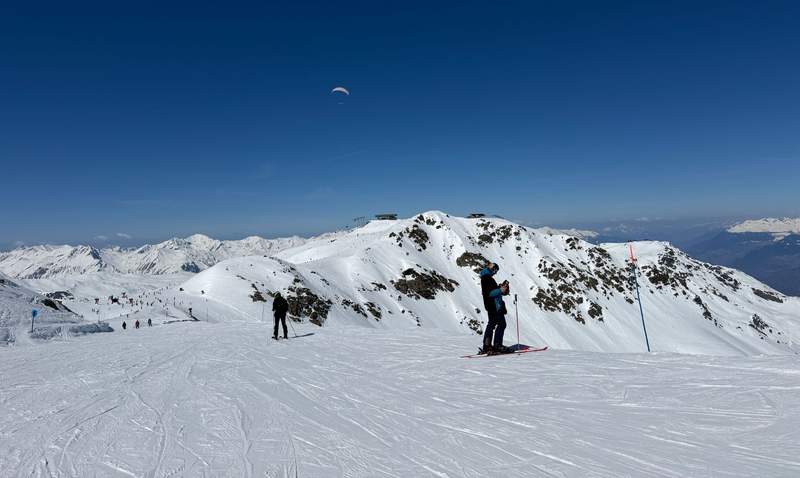 Skiers pause on groomed runs above Val Thorens near Caron 3200 with a paraglider soaring overhead and sweeping French Alps views under a deep blue sky.