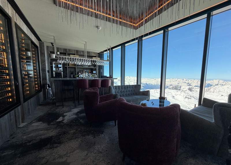Interior of Caron 3200 wine bar at Val Thorens with velvet armchairs wine wall and floor-to-ceiling windows overlooking snow-covered French Alps peaks.