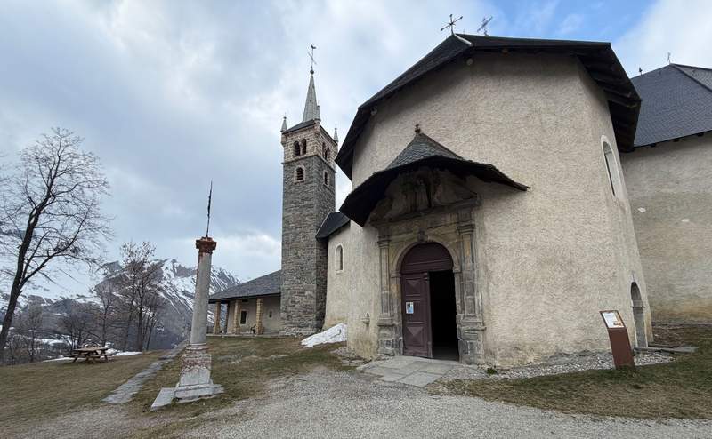 Notre Dame de la Vie Chapel in St Martin de Belleville with stone bell tower, an ornate doorway and snow-dusted Alpine peaks visible in the background.