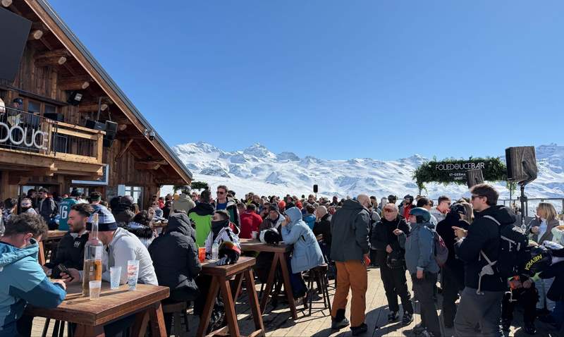 Crowded outdoor terrace at La Folie Douce Val Thorens with apres ski revelers wooden chalet building and snow covered French Alps peaks under a clear blue sky.