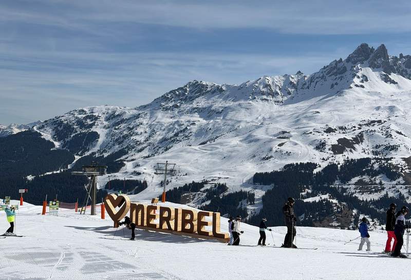 Skiers gathered around a large wooden Meribel sign on a groomed ski run with forested slopes and dramatic rocky peaks of Les 3 Vallees French Alps in the background.
