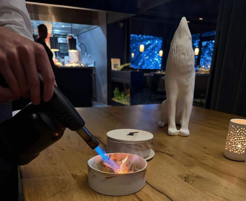 Chef torching pine needles tableside at Alpage restaurant inside Hotel Annapurna Courchevel in Les 3 Vallees with dramatic moody dining room in the background.