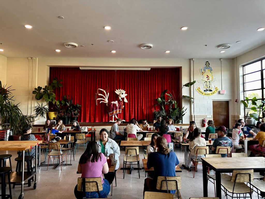 Interior of Feather Bone Provisions & Assembly restaurant in a repurposed elementary school auditorium with stage and school chairs.
